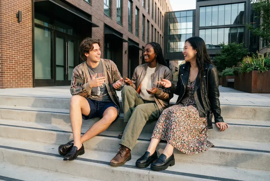 Three young friends sitting on concrete steps outside a modern building, wearing eclectic anti-fluency fashion including a vintage cardigan, silky bomber jacket, and prairie dress mixed with chunky boots and sneakers.