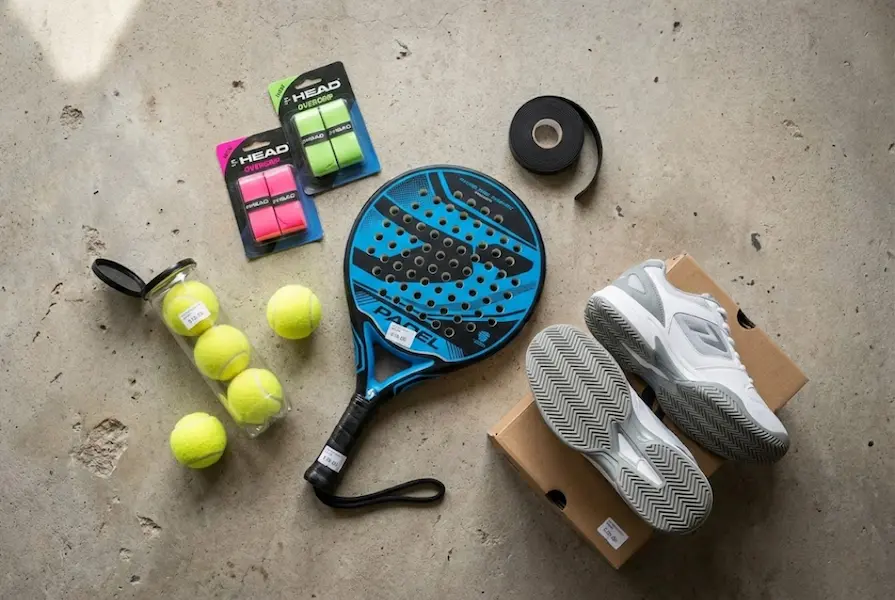 Overhead flat lay of essential padel equipment on a concrete floor, including a round-shaped racket, court shoes, a tube of yellow balls, and colorful overgrips.