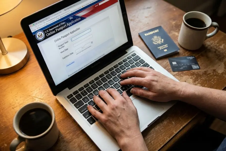 Overhead view of hands typing on a laptop displaying the official U.S. Department of State online passport application form, with a physical US passport book and credit card on a wooden desk.