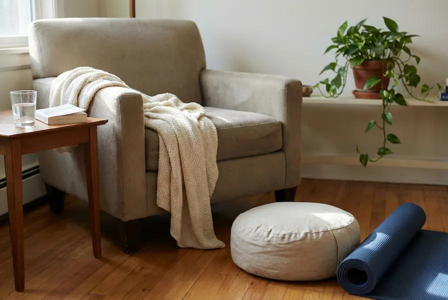 A calming home corner setup featuring a beige armchair with a knitted throw, a yoga mat, and a meditation cushion arranged for relaxation and yoga in soft natural light.