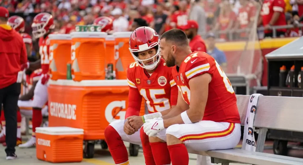 Quarterback Patrick Mahomes and tight end Travis Kelce sitting on the bench discussing game strategy while wearing Kansas City Chiefs uniforms.