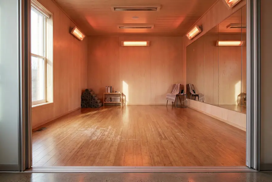 Interior view of an empty modern fitness studio featuring wall-mounted infrared heating panels, natural wooden flooring, large mirrors, and yoga equipment in a warm, minimalist setting.