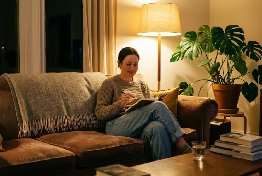 A person sitting comfortably on a living room couch under warm lamp light, holding a pen and notebook for evening journaling, surrounded by cozy decor including a throw blanket and plants.