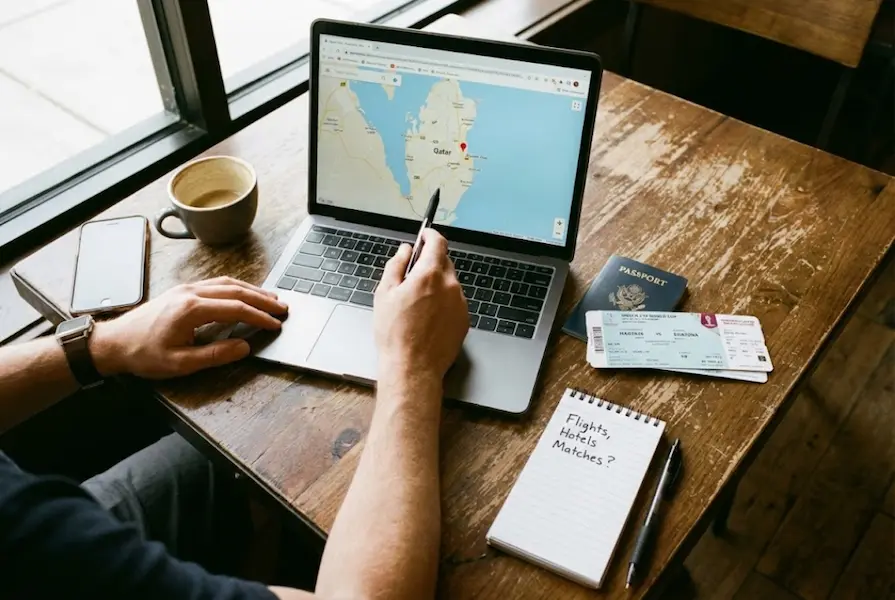Overhead view of a person planning a trip at a wooden coffee shop table, featuring an open laptop displaying a map, a passport, coffee, and scattered World Cup match tickets.