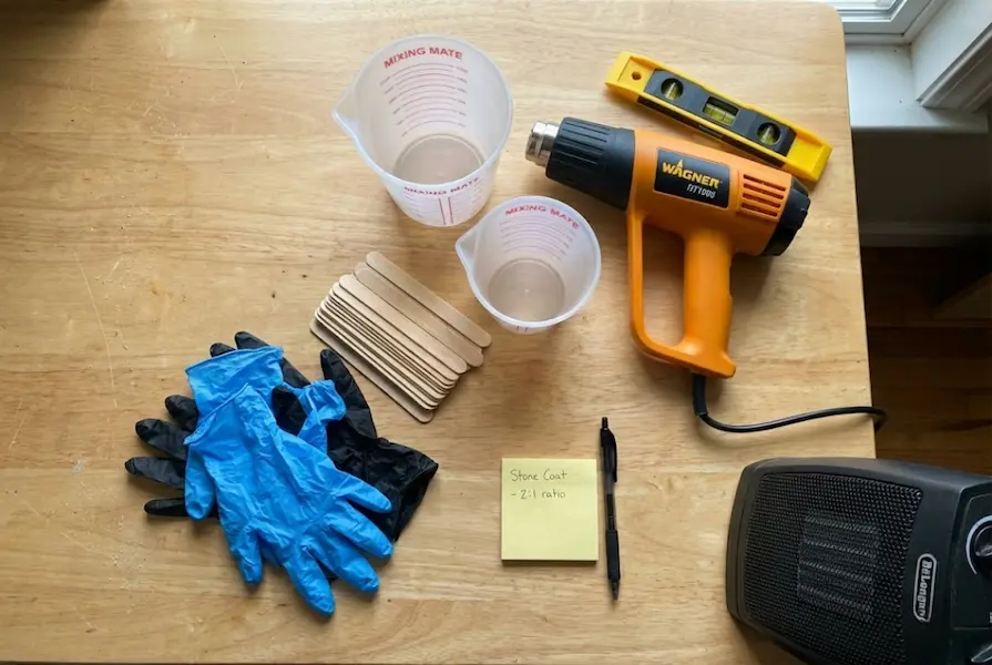 Flat lay on a wooden table showing resin art supplies including a Wagner heat gun, silicone measuring cups, nitrile gloves, and a handwritten mixing ratio note.