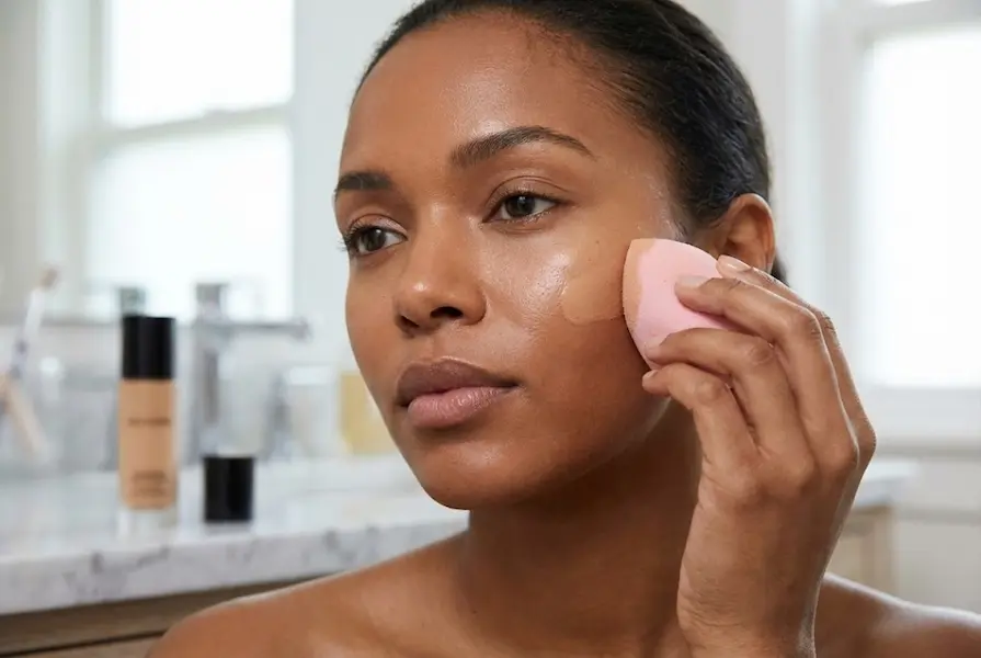 Close-up of a woman with medium-dark skin bouncing a damp pink beauty sponge on her cheek to blend foundation, showing a smooth, healthy skin texture with a blurred foundation bottle in the background.
