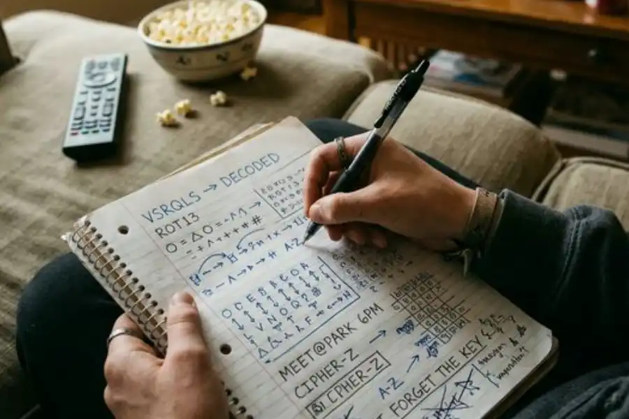 A teenager's hands holding a pen over a notebook filled with handwritten coded messages and cipher work, with a TV remote and popcorn nearby on a couch